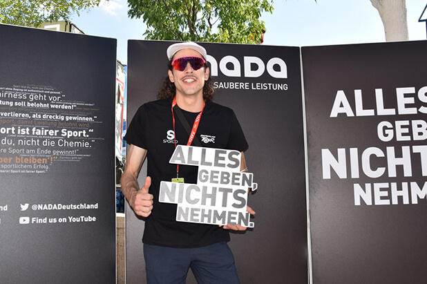 Radsportfan mit Statement-Schild vor "Alles geben nichts nehmen"-Wand