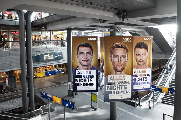 Drei große Werbeplakate mit Porträts von Klitschko, Reckermann und Popow und dem Text 'ALLES GEBEN, NICHTS NEHMEN' in einem modernen Bahnhof mit Rolltreppen und Geschäften im Hintergrund