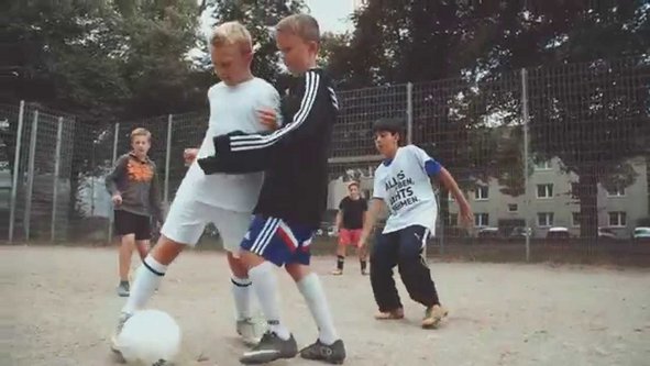 Zwei Jungen kämpfen um einen Fußball auf einem staubigen Spielfeld, weitere Kinder beobachten im Hintergrund.
