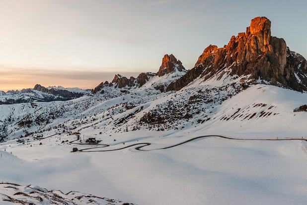Verschneite Berglandschaft bei Sonnenaufgang mit schroffen Felsen und einer kurvigen Straße.